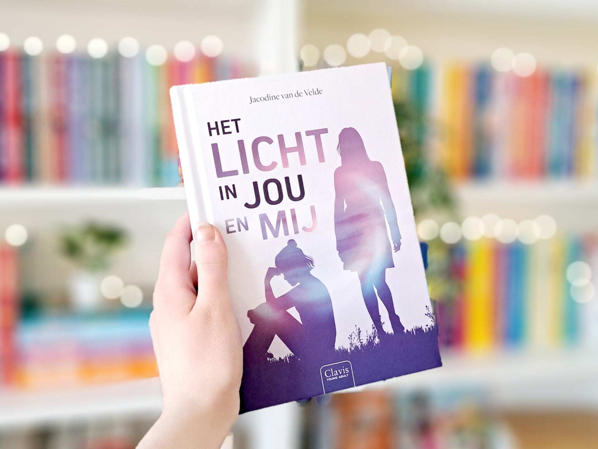 Een witte hand houdt 'het licht in jou en mij' van Jacodine van de Velde omhoog tegen een wazige boekenkast met lichtjes. Foto bij recensie door turnthepages.nl Rolstoelgebruiker en disabled bookstagrammer en blogger.