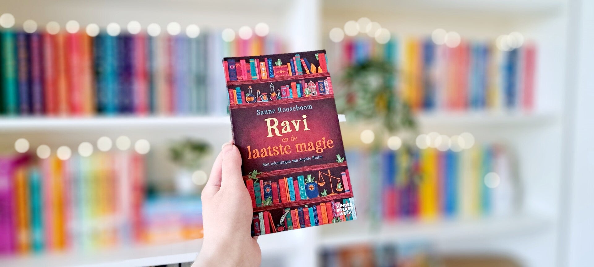 Een witte hand houdt 'Ravi en de laatste magie' van Sanne Rooseboom omhoog voor een kleurrijke wazige boekenkast met lichtjes. Foto bij own-voices recensie door turnthepages.nl Rolstoelgebruiekr en disabled bookstagrammer en blogger.