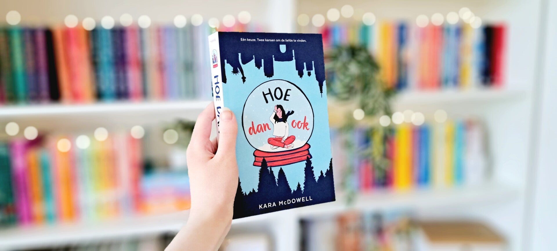 Een witte hand houdt 'Hoe dan ook' van Kara McDowell omhoog voor een kleurrijke wazige boekenkast met lichtjes. Foto bij recensie door turnthepages.nl Rolstoelgebruiker, disabled bookstagrammer en queer blogger.