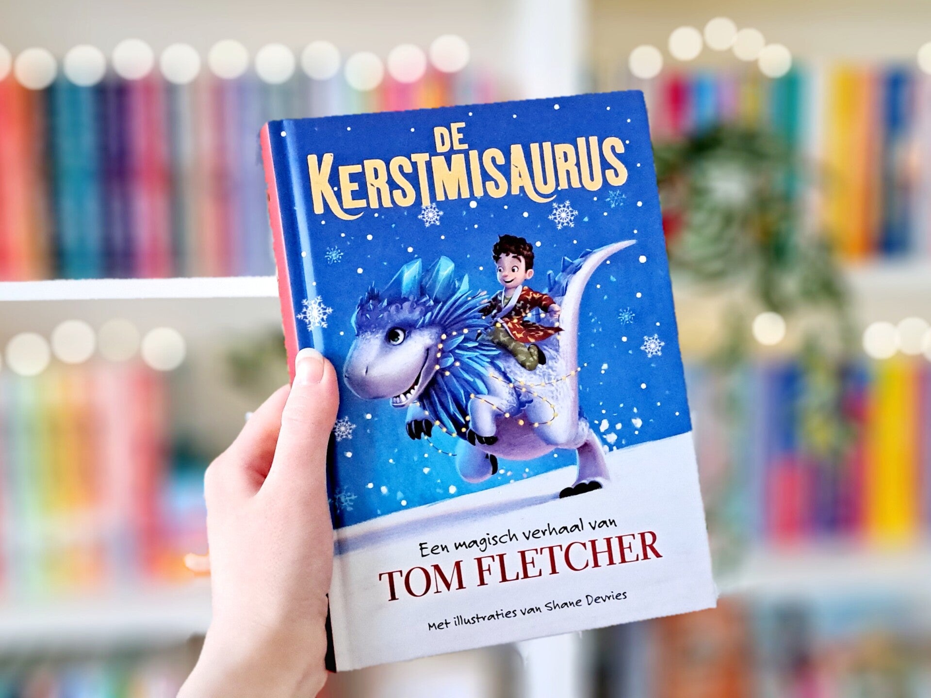 Een witte hand houdt 'De kerstmisaurus' van Tom Fletcher omhoog voor een kleurrijke wazige boekenksat met lichtjes. Foto bij own-voices recensie door turnthepages.nl Rolstoelgebruiker en disabled bookstagrammer en blogger.