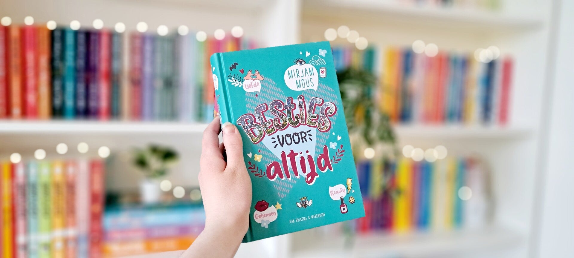 Een witte hand houdt 'Besties voor altijd' door Mirjam Mous omhoog voor een kleurrijke wazige boekenkast met lichtjes. Foto bij recensie door turnthepages.nl Rolstoelgebruiker en disabled bookstagrammer en blogger.
