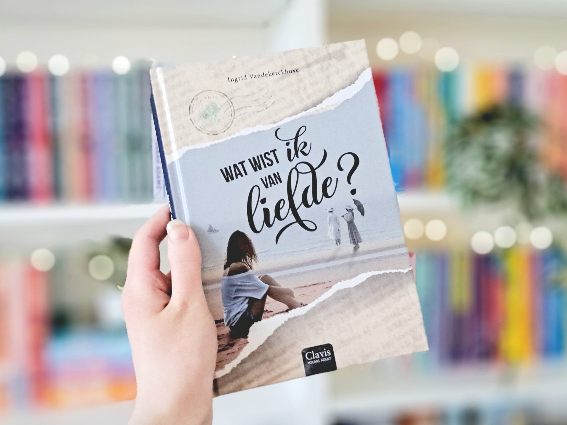 Een witte hand houdt 'Wat wist ik van liefde?' van Ingrid Vandekerckhove omhoog voor een kleurrijke wazige boekenkast met lichtjes. Foto bij recensie door turnthepages.nl Rolstoelgebruiker, disabled bookstagrammer en queer blogger.