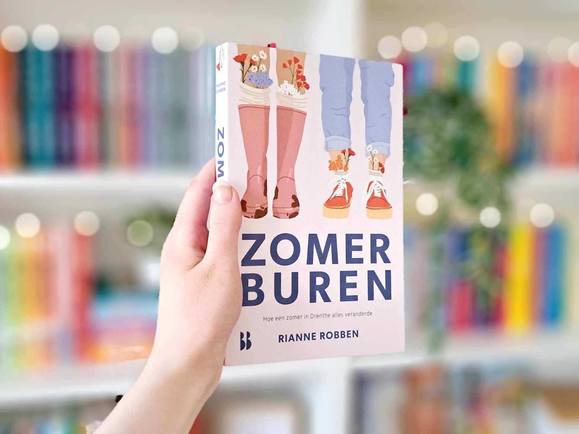 Een witte hand houdt 'Zomerburen' van Rianne Robben omhoog voor een kleurrijke wazige boekenkast met lichtjes. Foto bij recensie door turnthepages.nl Rolstoelgebruiker en  disabled bookstagrammer en queer blogger met ADHD.