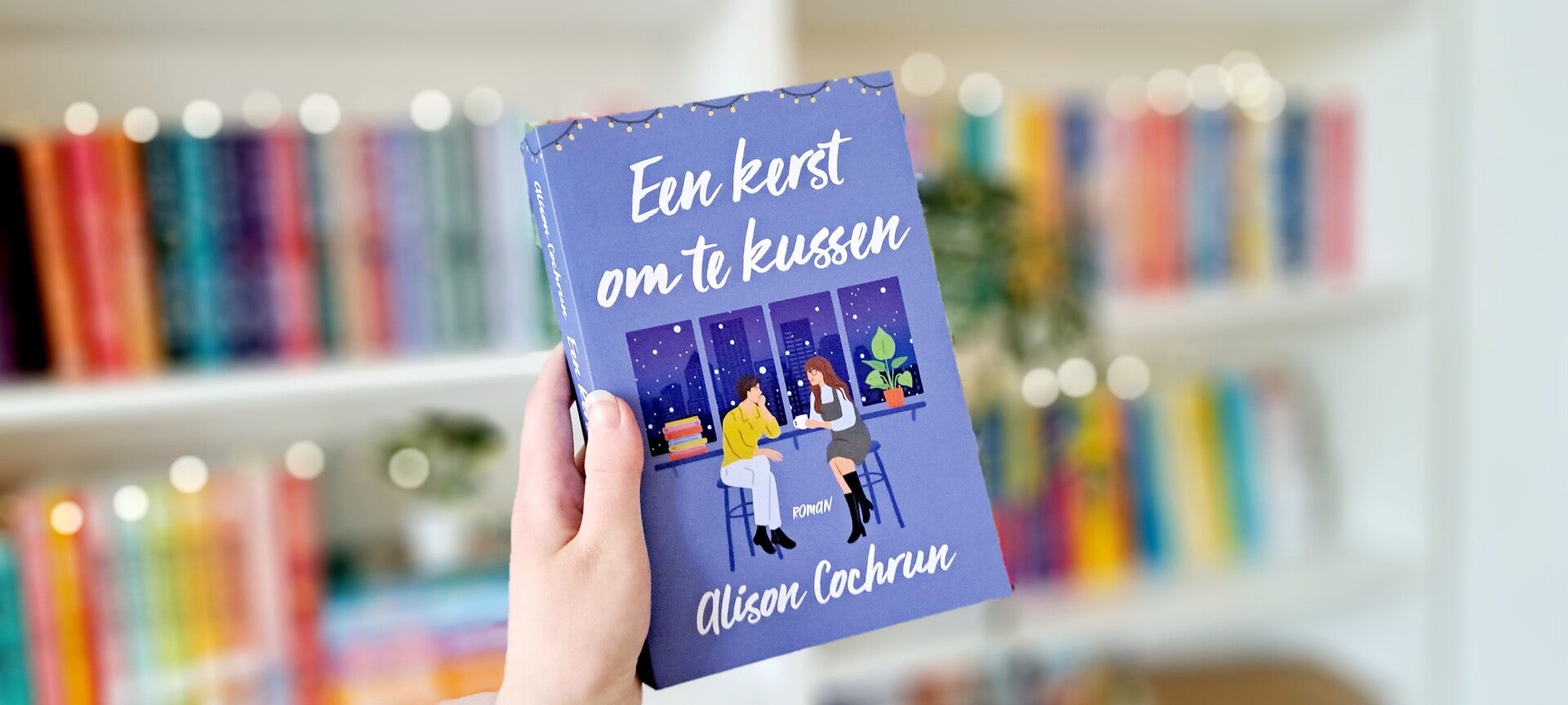 Een witte hand houdt 'Een kerst om te kussen' van Alison Cochrun omhoog voor een kleurrijke wazige boekenkast met lichtjes. Foto bij recensie door turnthepages.nl Rolstoelgebruiker en disabled bookstagrammer en blogger met ADHD./