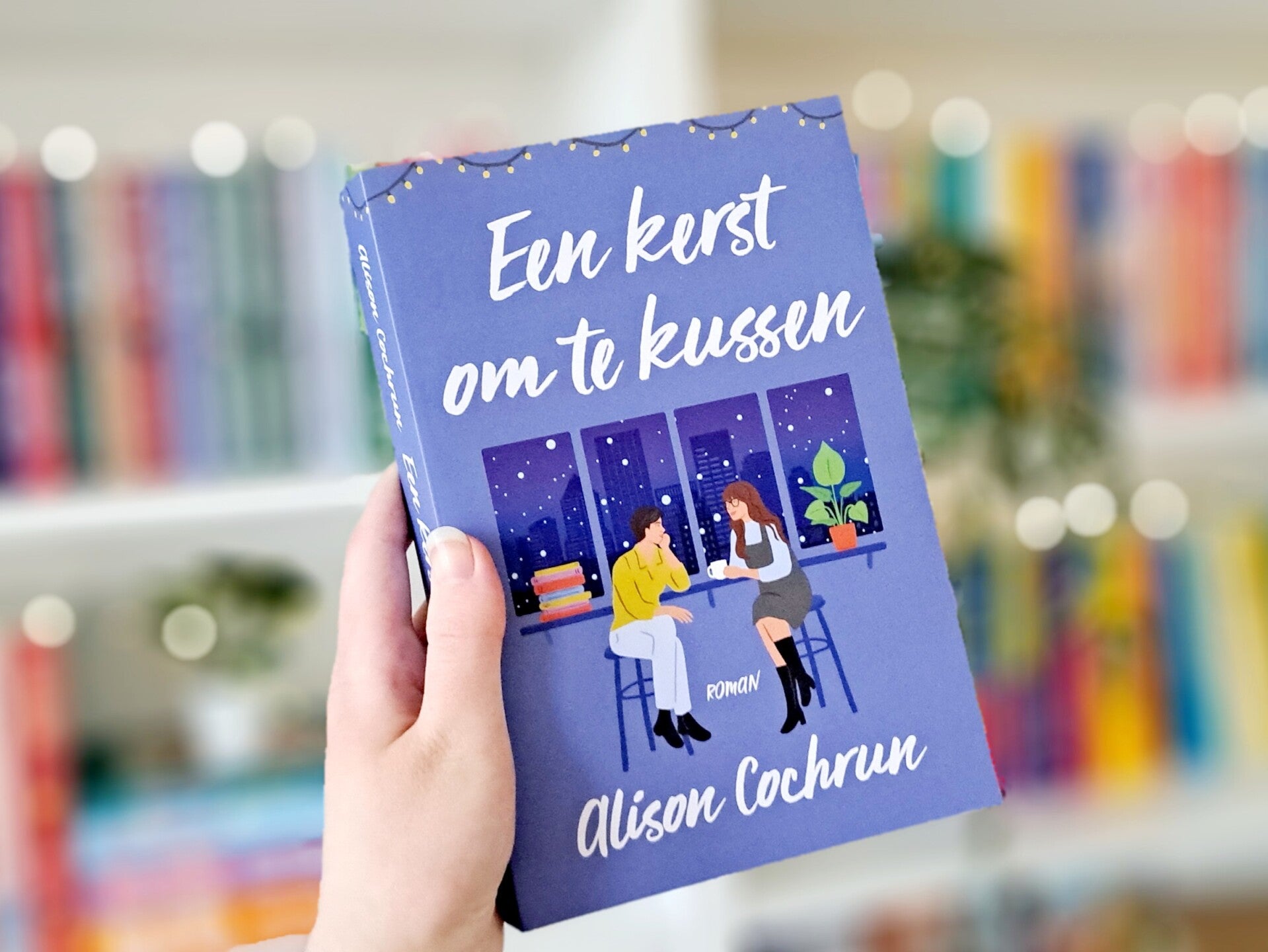 Een witte hand houdt 'Een kerst om te kussen' van Alison Cochrun omhoog voor een kleurrijke wazige boekenkast met lichtjes. Foto bij recensie door turnthepages.nl Rolstoelgebruiker en disabled bookstagrammer en blogger met ADHD./