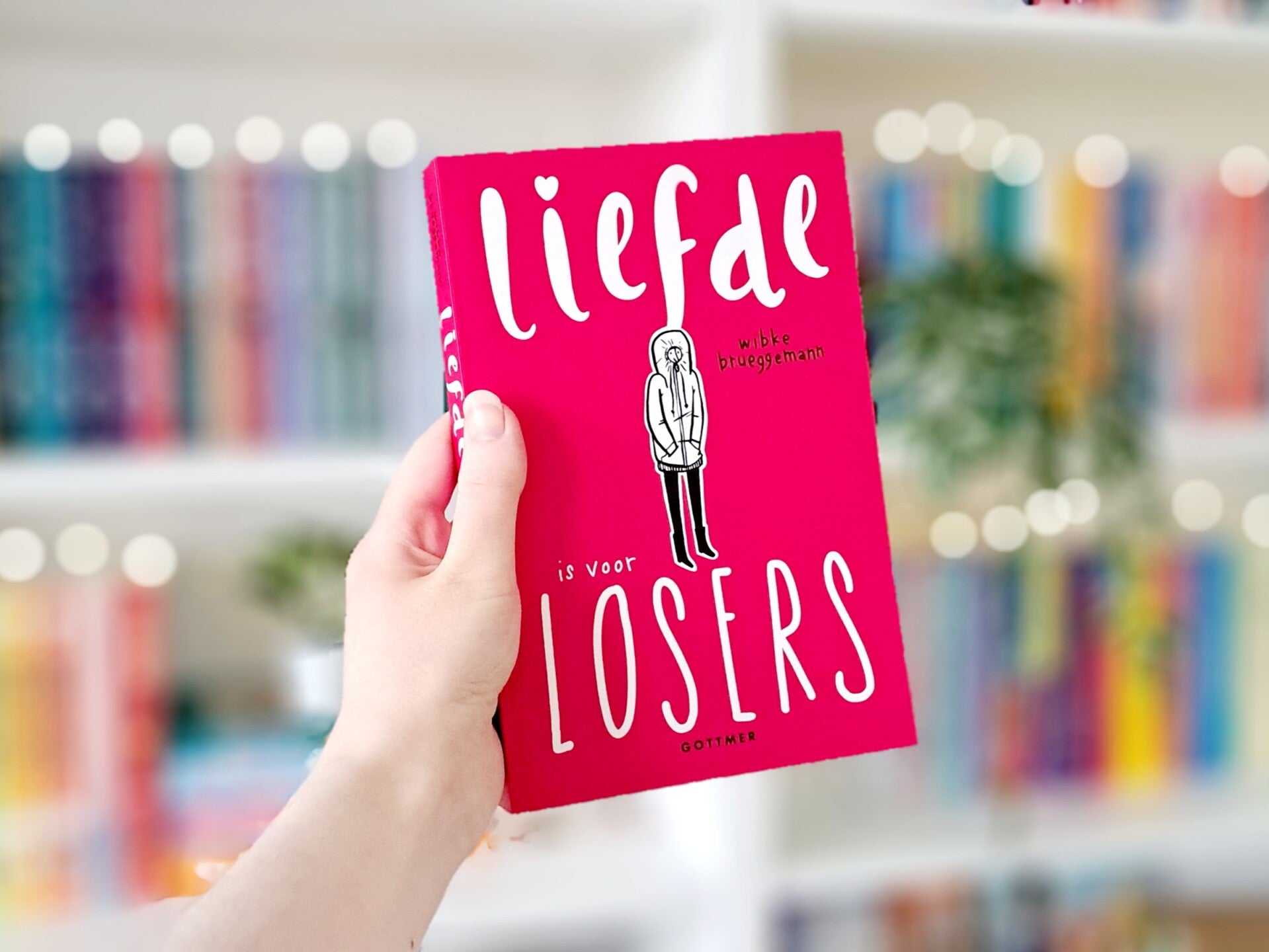 Een witte hand houdt 'Liefde is voor losers' van Wibke Brueggemann omhoog voor een kleurrijke wazige boekenkast met lichtjes. Foto bij recensie door turnthepages.nl Rolstoelgebruiker en disabled bookstgrammer en blogger.
