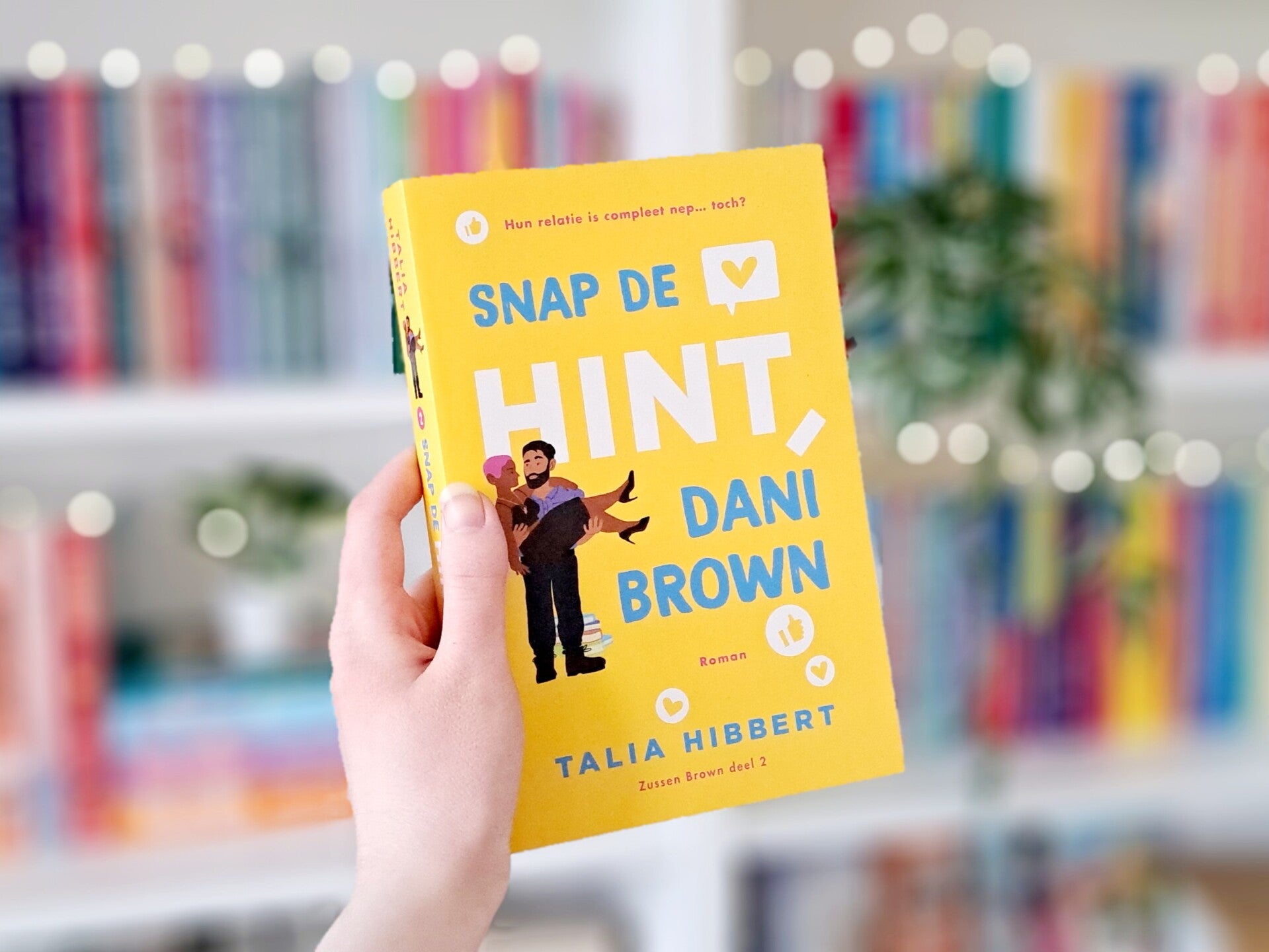 Een witte hand houdt 'Snap de hint, Dani Brown' deel twee van de Brown Sisters trilogie van Talia Hibbert omhoog voor een kleurrijke wazige boekenkast met lichtjes. Foto bij recensie door turnthepages.nl Rolstoelgebruiker, disabled bookstagrammer en queer
