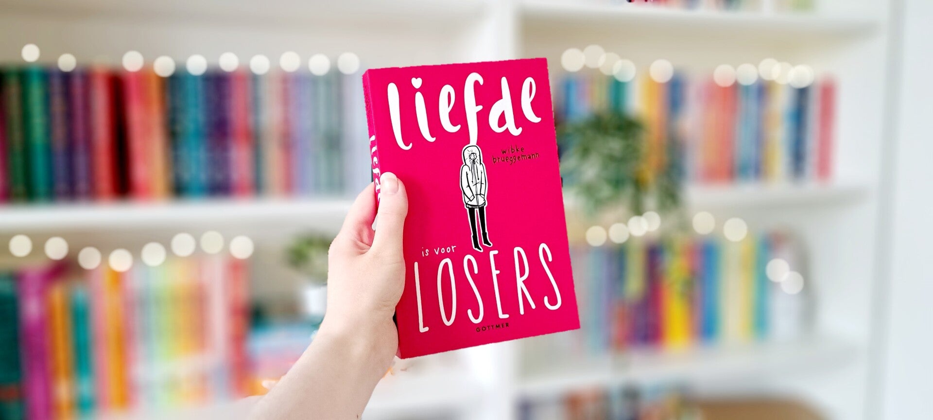 Een witte hand houdt 'Liefde is voor losers' van Wibke Brueggemann omhoog voor een kleurrijke wazige boekenkast met lichtjes. Foto bij recensie door turnthepages.nl Rolstoelgebruiker en disabled bookstgrammer en blogger.
