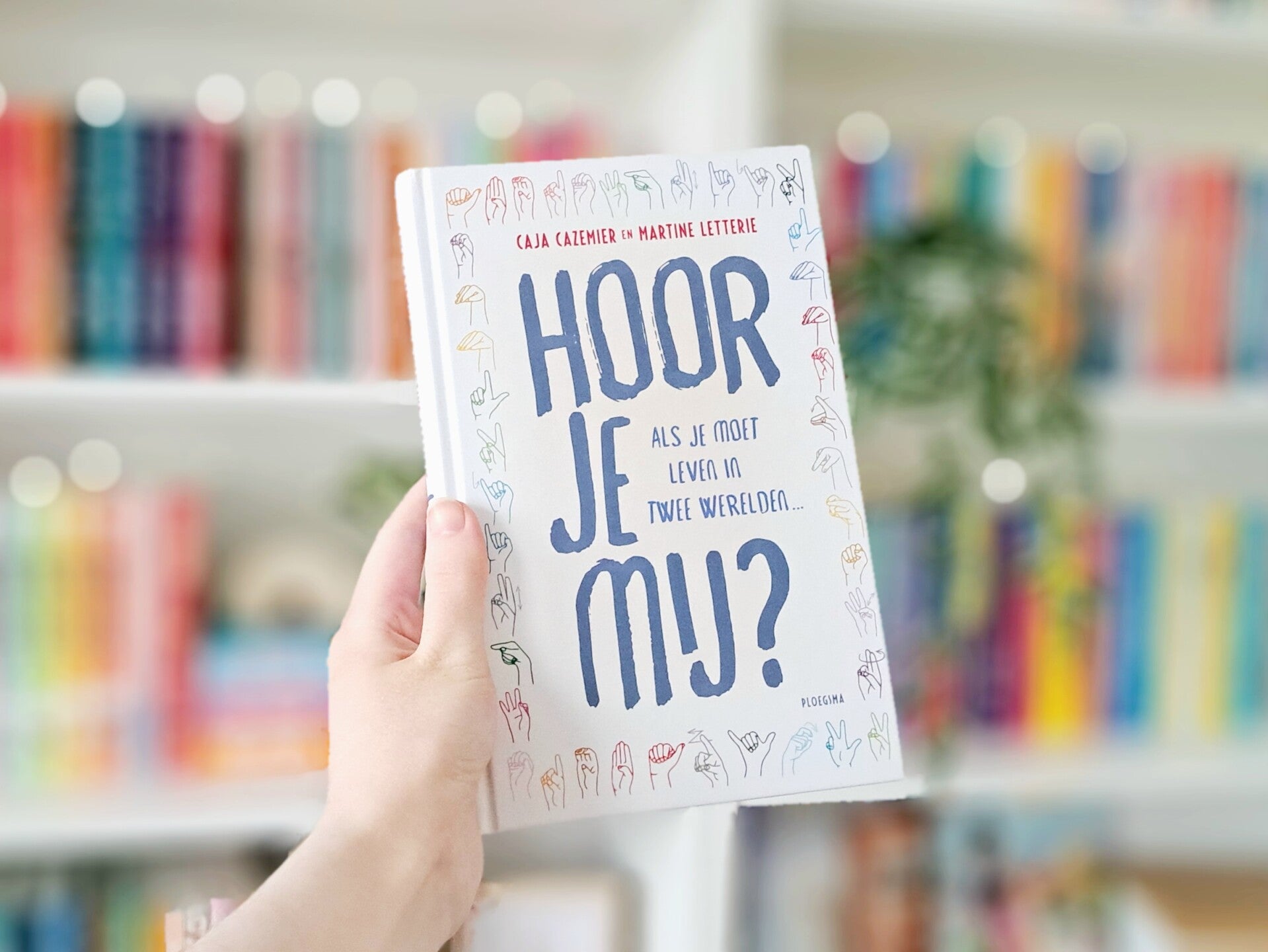 Een witte hand houdt 'Hoor je mij?' van Caja Cazemier en Martine Letterie omhoog voor een kleurrijke wazige boekenkast met lichtjes. Foto bij recensie door turnthepages.nl | Disabled bookstagrammer en boekblogger