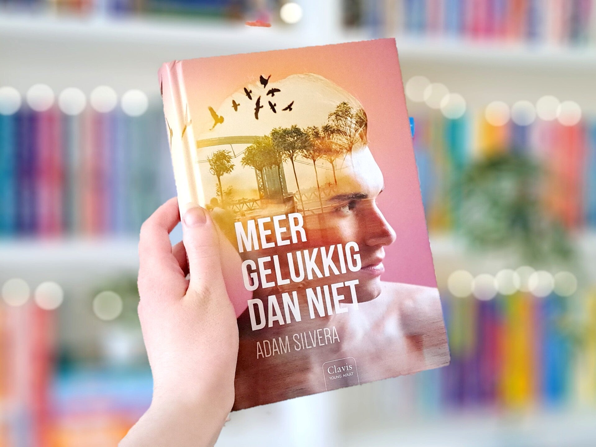 Een witte hand houdt 'Meer gelukkig dan niet' van Adam Silvera omhoog voor een kleurrijke wazige boekenkast met lichtjes Foto bij recensie door turnthepages.nl