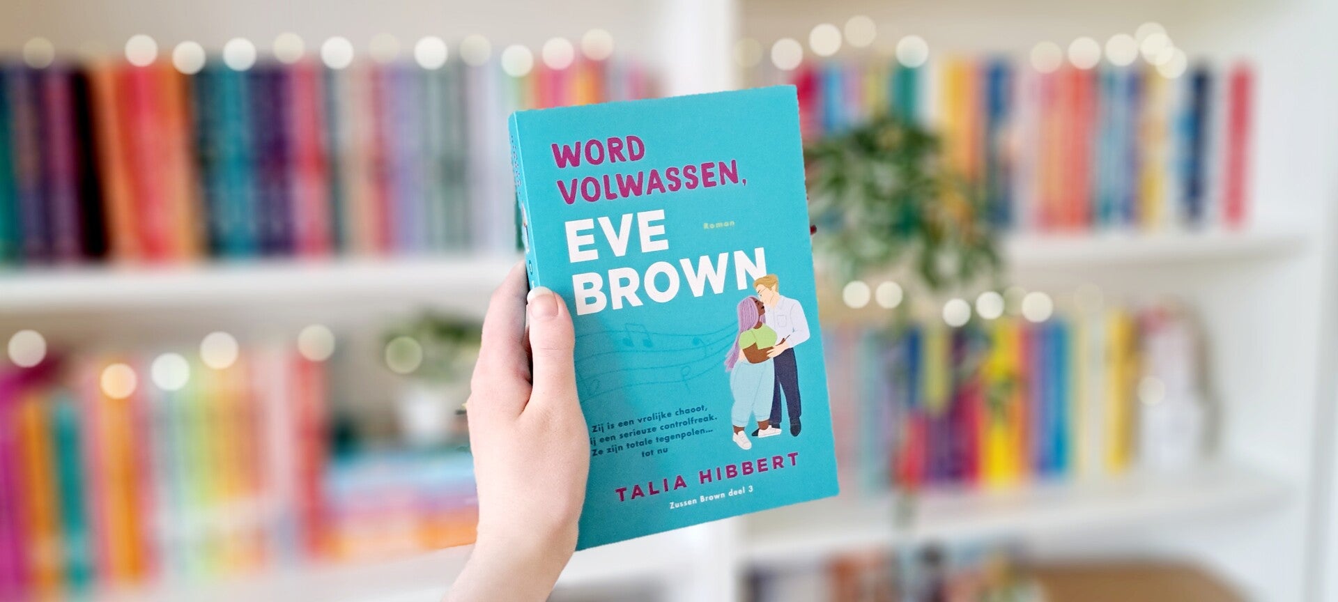 Een witte hand houdt 'Word volwassen, Eve Brown' deel drie in the sisters brown trilogie van Talia Hibbert omhoog voor een kleurrijke wazige boekenkast met lichtjes. Foto bij recensie door turnthepages.nl Rolstoelgebruiker en disabled bookstagrammer en bl