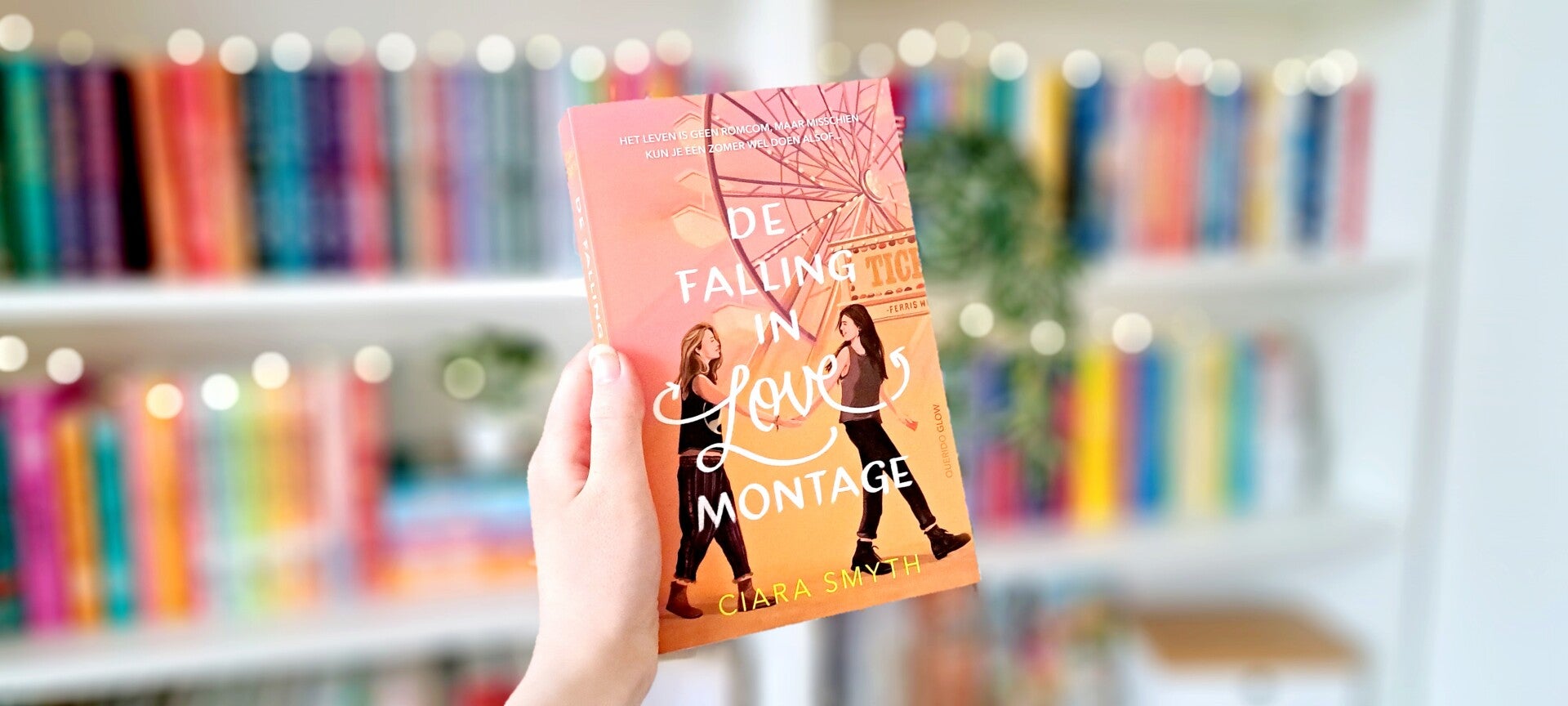 Een witte hand houdt 'De falling in love montage' van Ciara Smyth omhoog voor een kleurrijke wazige boekenkast met lichtjes. Foto bij recensie door turnthepages.nl Rolstoelgebruiker en disabled bookstagrammer en blogger.