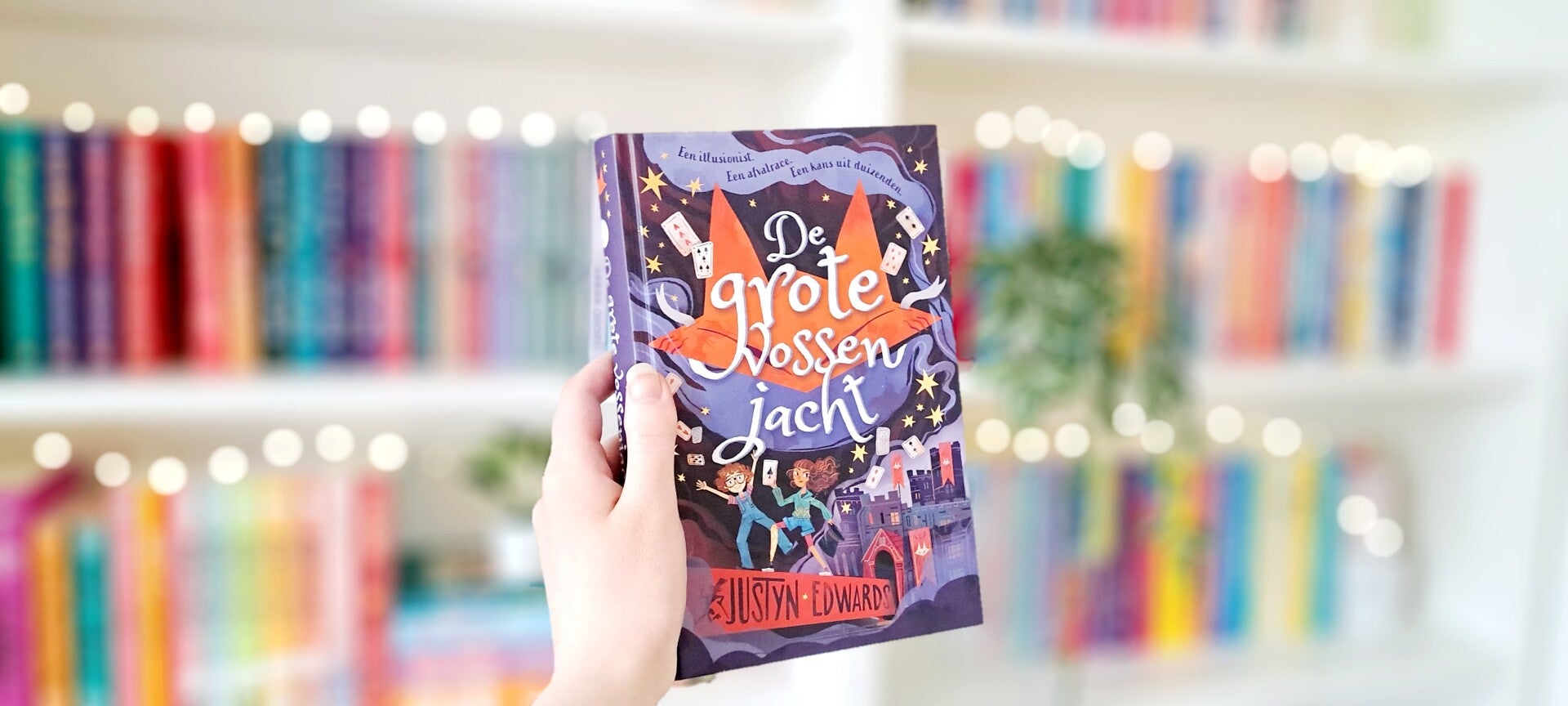 Een witte hand houdt 'De grote vossenjacht' van Justyn Edwards omhoog voor een kleurrijke wazige boekenkast met lichtjes. Foto bij recensie door turnthepages.nl Rolstoelgebruiker en disabled bookstagrammer en blogger.