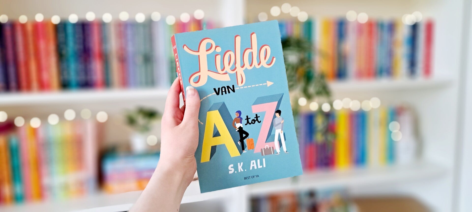Een witte hand houdt 'Liefde van A tot Z' van S.K. Ali omhoog voor een kleurrijke wazige boekenkast met lichtjes. Foto bij recensie door turnthepages.nl Rolstoelgebruiker en disabled bookstagrammer en blogger.
