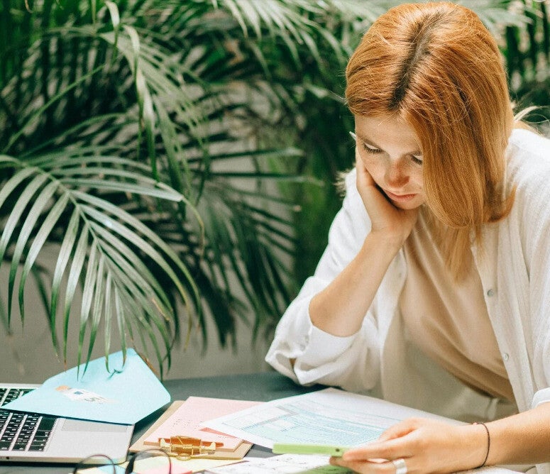 Frau konzentriert bei der Arbeit mit Dokumenten und Laptop an einem Schreibtisch mit grünen Pflanzen, um die Vereinfachung von Marketing-Komplexität zu illustrieren.