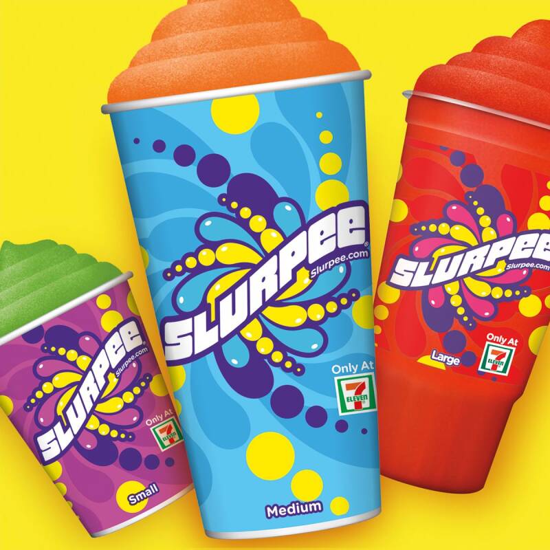 A colorful Slurpee in a clear cup with the 7-Eleven logo, sitting on a counter with bright summer lighting.
