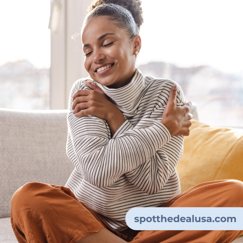A woman sitting comfortably on a couch, smiling gently as she wraps her arms around herself in a self-soothing, relaxing hug — a peaceful moment of self-care.