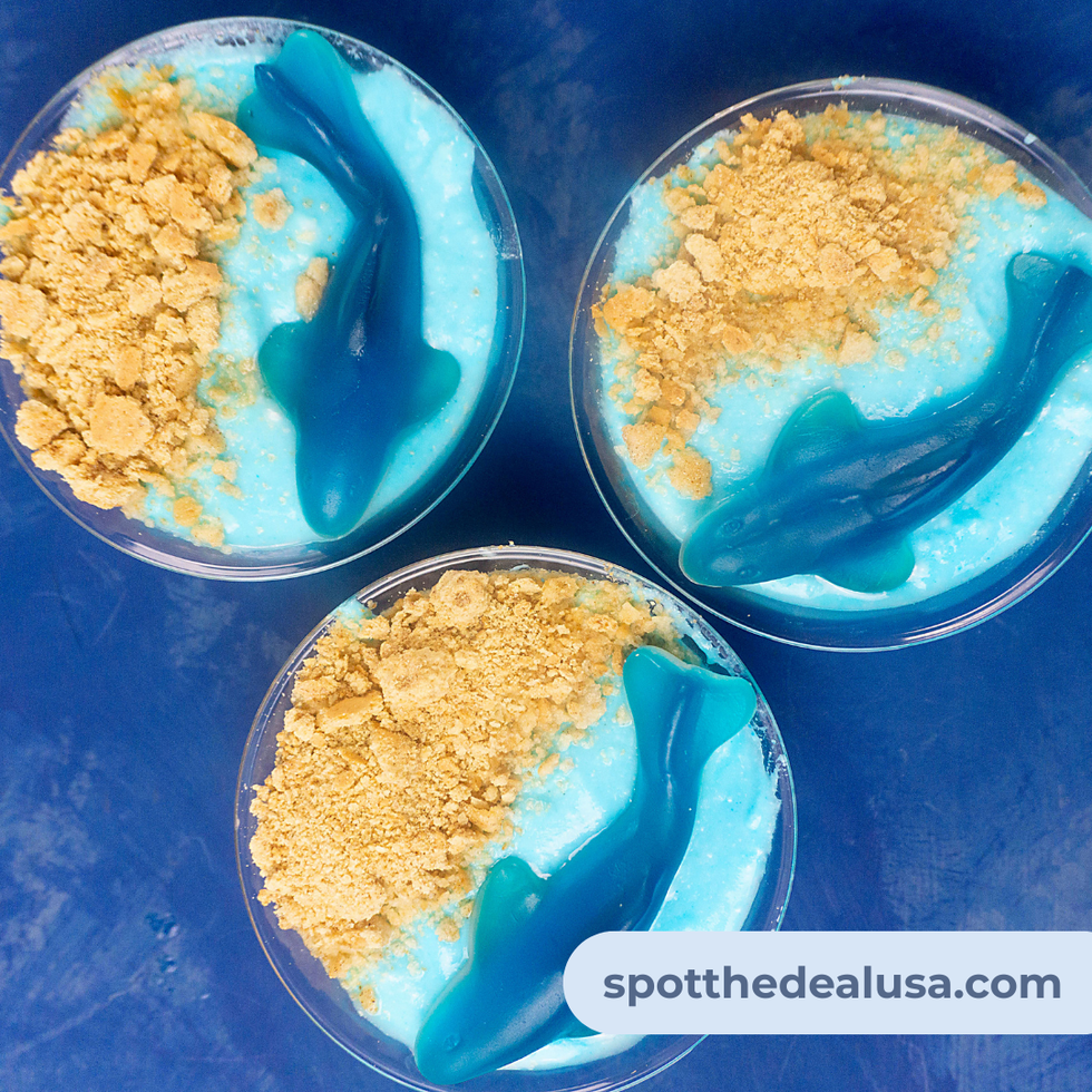 A clear plastic cup layered with ocean-blue yogurt, topped with crushed graham crackers and a single shark gummy. A fun and kid-friendly beach snack for summer or Shark Week.