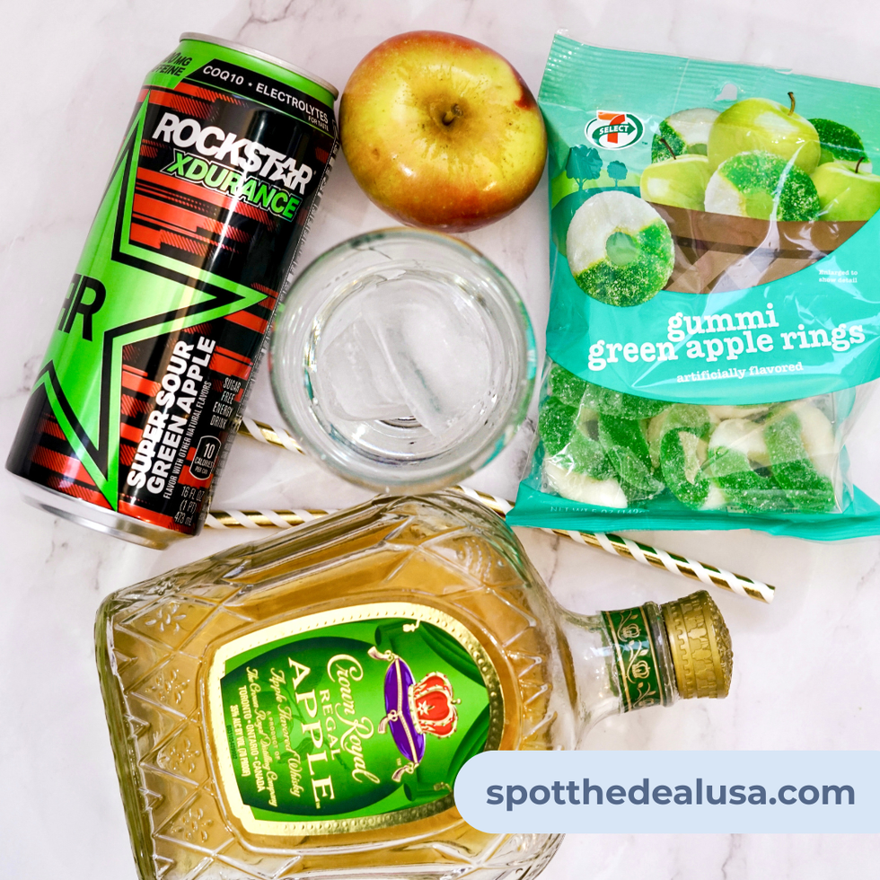 Ingredients for Sparkling Apple Buzz cocktail including apple-flavored whiskey, sour green apple energy drink, green apple gummy rings, and ice cubes displayed on a countertop.