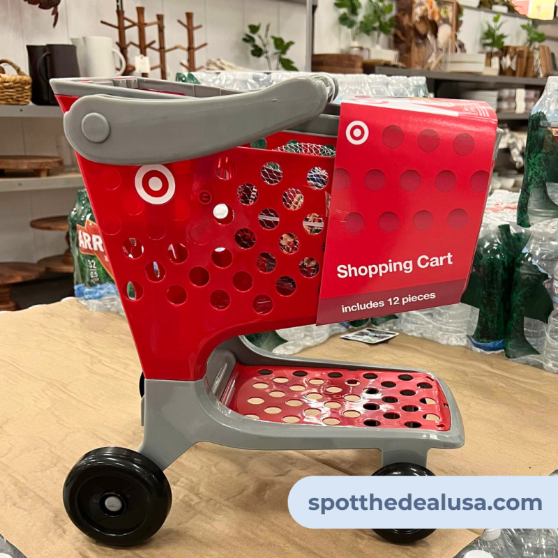 Target toy shopping cart with red plastic frame, filled with mini grocery items and a doll seat, placed on a hardwood floor in a cozy home setting.