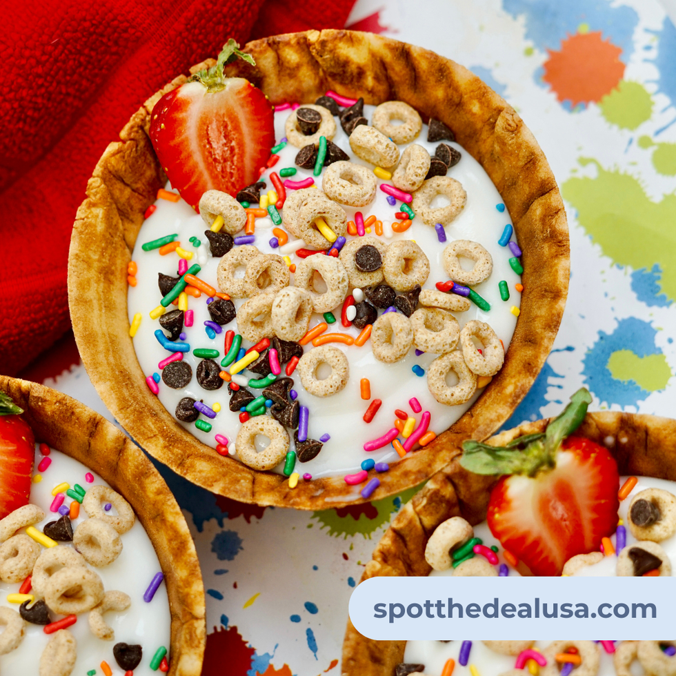A colorful yogurt sundae bowl in a waffle cone, topped with Cheerios, chocolate chips, sprinkles, and sliced strawberries.