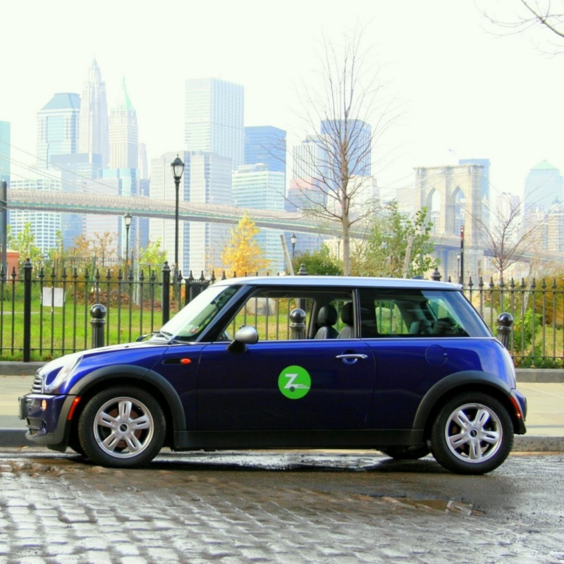 Blue Mini Cooper Zipcar parked with a view of the Boston skyline, Zipcar logo visible on driver side door.