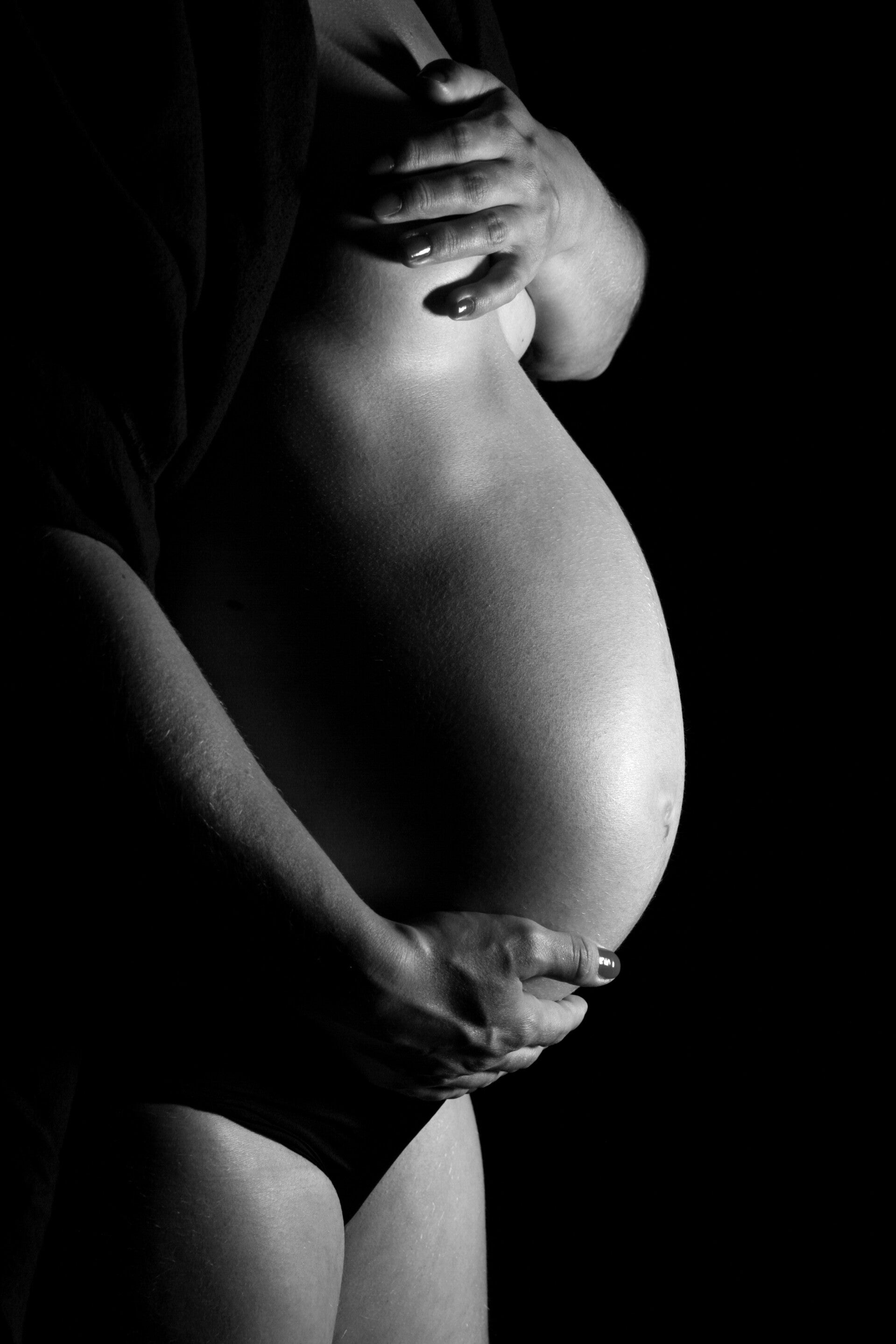 Schwarz-Weiß-Aufnahme eines Babybauchs in der Schwangerschaft vor dunklem Hintergrund.