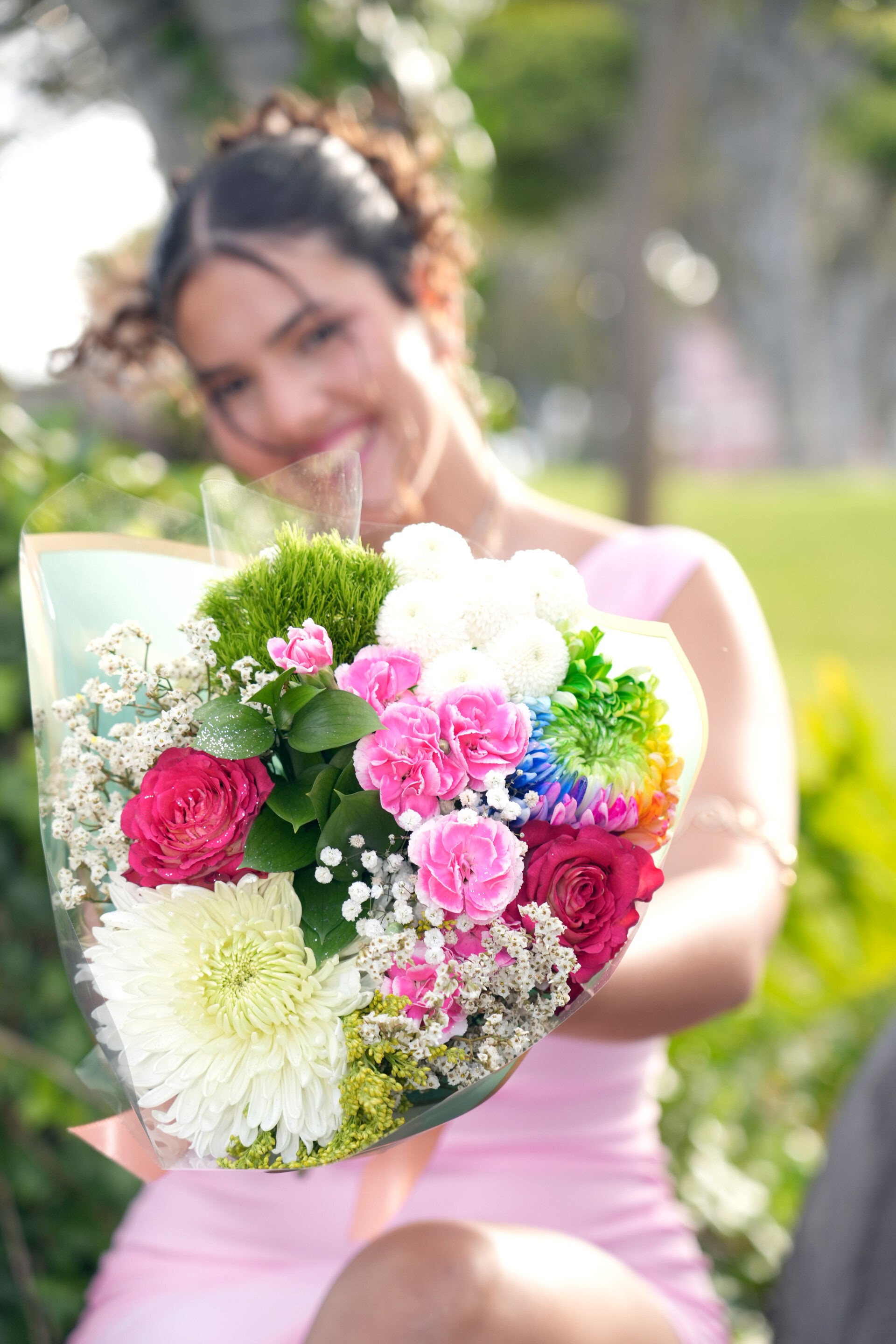 Junge Frau lächelt strahlend im Park und hält einen großen, bunten Blumenstrauß im Arm.