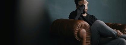 Man sitting on a brown leather armchair with his legs crossed, resting his forehead in his hand, he subdued setting suggests a quiet moment of reflection or seeking clarity in a calm space.