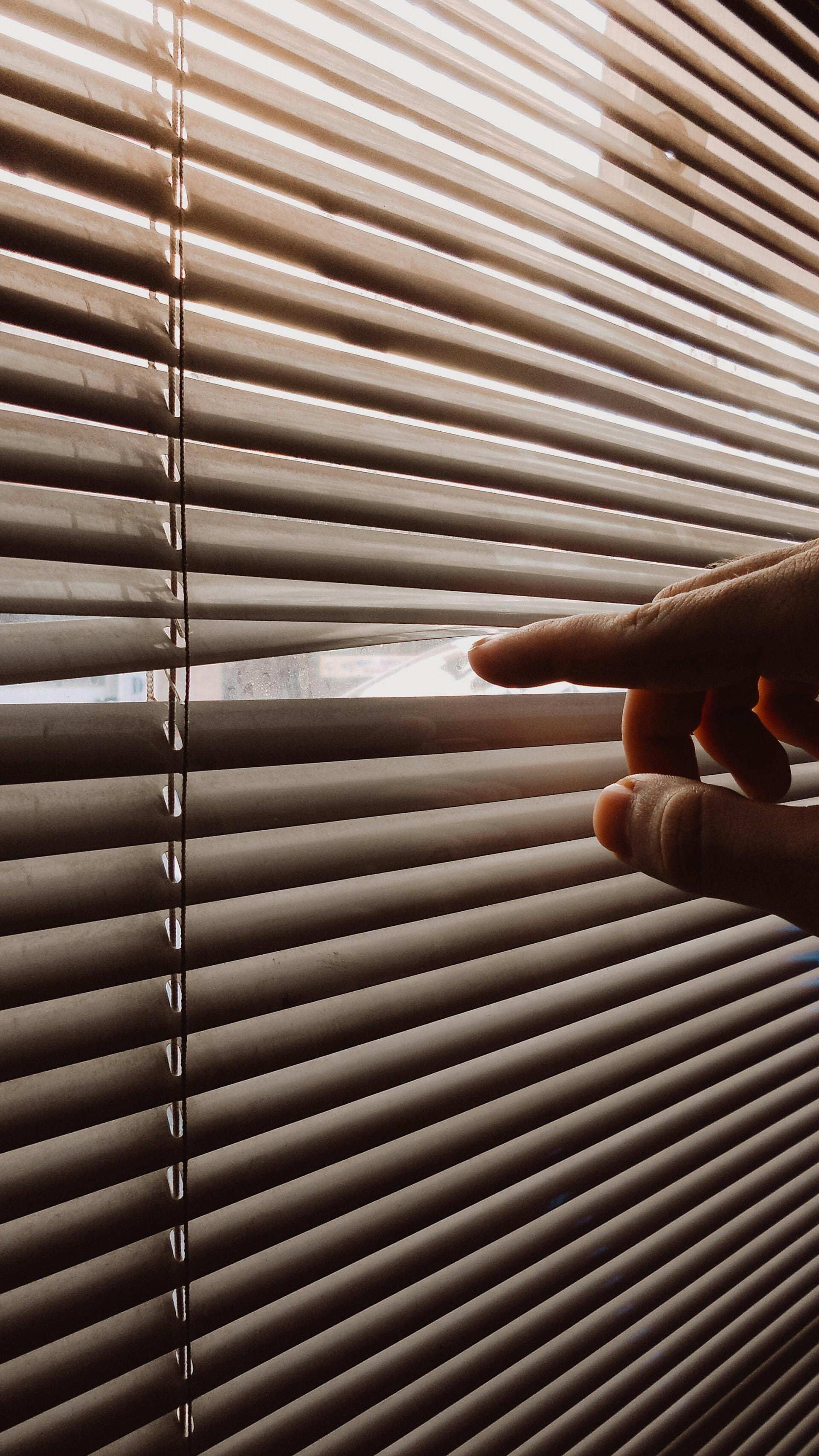 Image of closed venitian blinds  with a persons hand  using their index finger to slightly part the blinds.