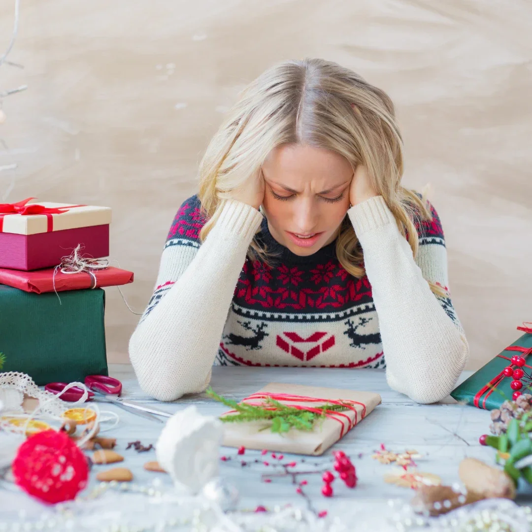 A women sitting at a table  wearing a Christmas Jumper in the process of wrapping presents  but with her head in her hands as if in dispair or overwhelmed