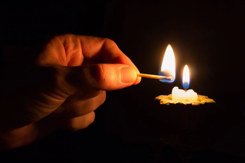 Image of a persons hand holding a lit match against a lit candle, against a black colour background