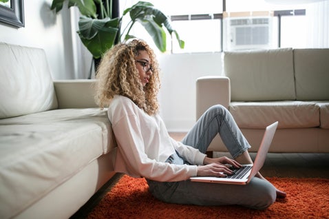 Woman with curly hair and glasses sitting on an orange rug, leaning against a sofa and using a laptop during a virtual therapy session in a bright living room.