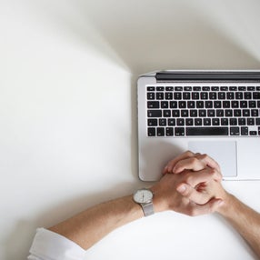 Aerial view of a man with clasped hands resting on a white desk, beside an open laptop, suggesting a moment of pause before or during an online counselling or therapy session.