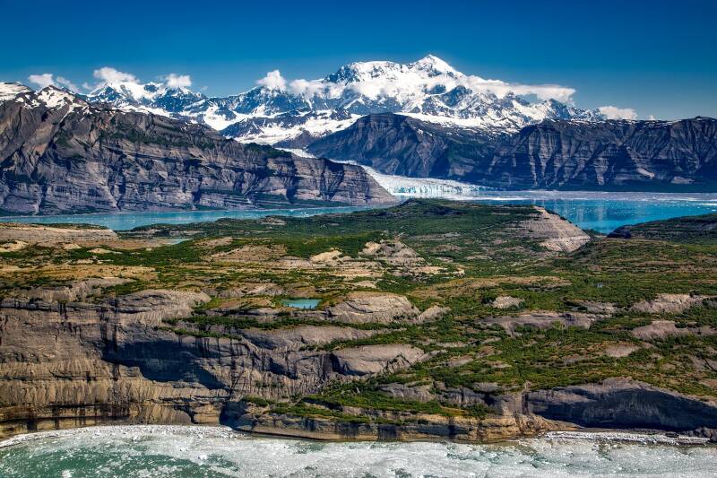 Image of Alaska showcasing a breathtaking view of snowy mountains reflected in crystal clear water under a clear blue sky.