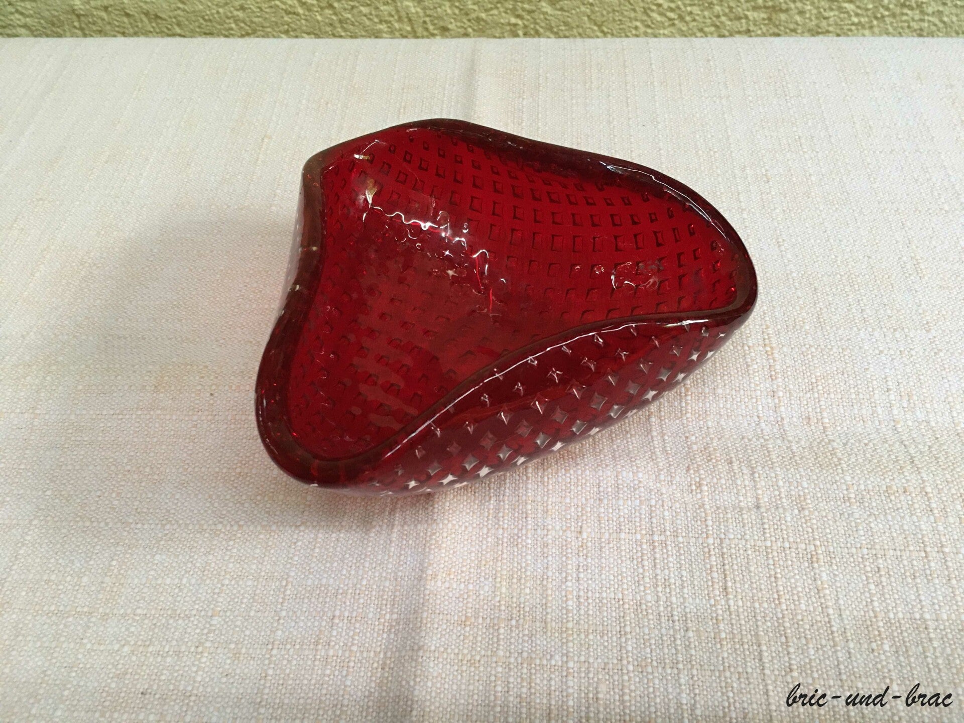 Der Aschenbecher ist aus Glas in gewölbter Form mit Einschlüssen und 3 nach innen eingefalteten Rändern in der Mitte, die als Zigarettenablagen dienen. Der Boden ist plan geschliffen