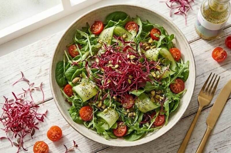 En skål fylld med en grön sallad med gurka och tomater, rikligt toppad med mörkröda amaranthskott odlade från egna röda amaranthfrön, placerad på ett träbord tillsammans med bestick.