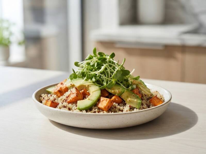 Närbild av en hälsosam Power Bowl i en skål på en ljus träbänk. Basen består av quinoa toppad med tärnad, rostad sötpotatis, skivad avokado och en generös hög med färska, gröna solrosskott. Köksmiljö i bakgrunden.