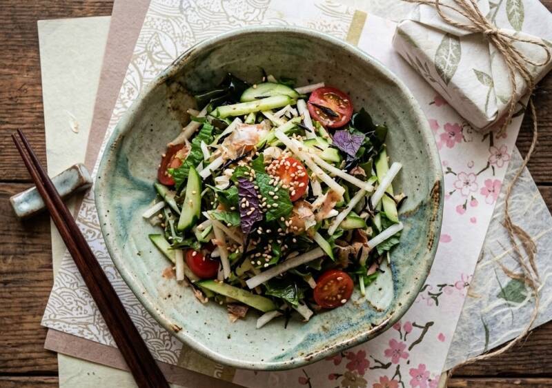 En japansk sallad i en rustik skål med gröna och röda shiso-skott, rättika, körsbärstomater och rostade sesamfrön, serverad på ett underlag av handgjort Washi-papper med ätpinnar bredvid.