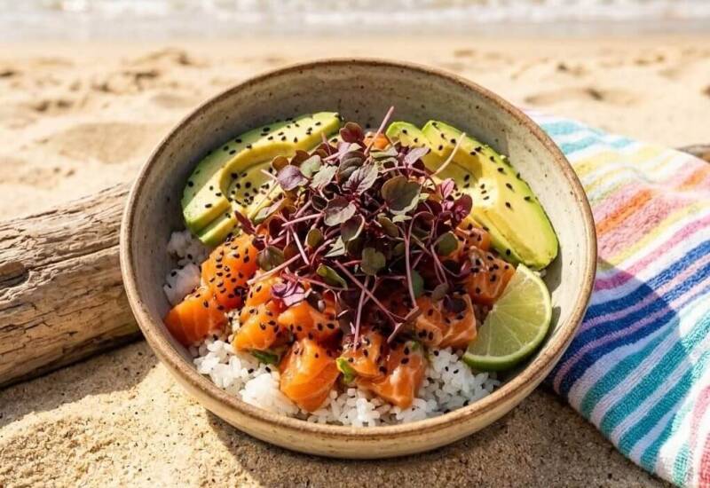 En skål med lax, ris, avokado och lime på en sandstrand, toppad med röda shisoskott odlade från egna röda shisofrö / perillafrö.