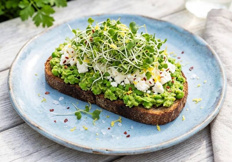 En rostad surdegsmacka med mosade ärtor och fetaost på en tallrik, toppad med broccoliskott och groddar odlade från egna broccolifrön. Calabrese