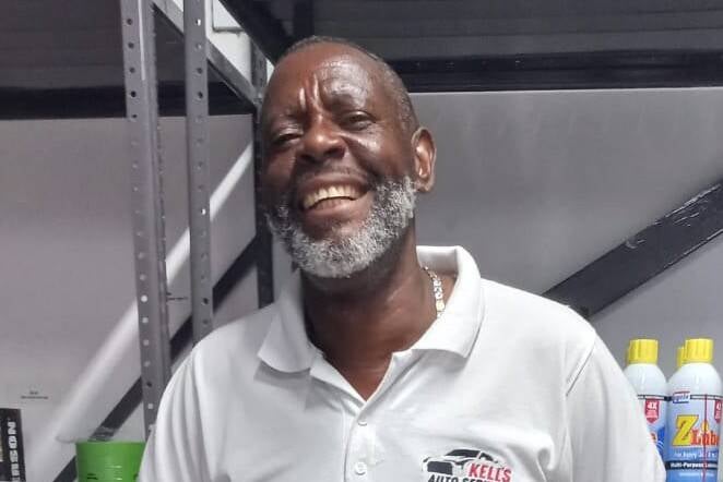 A smiling man in a white "KEL'S AUTO SERVICE" polo shirt and jeans stands in front of shelves stocked with automotive supplies, including oil containers, car polish, paint cans, and windshield wiper blades.