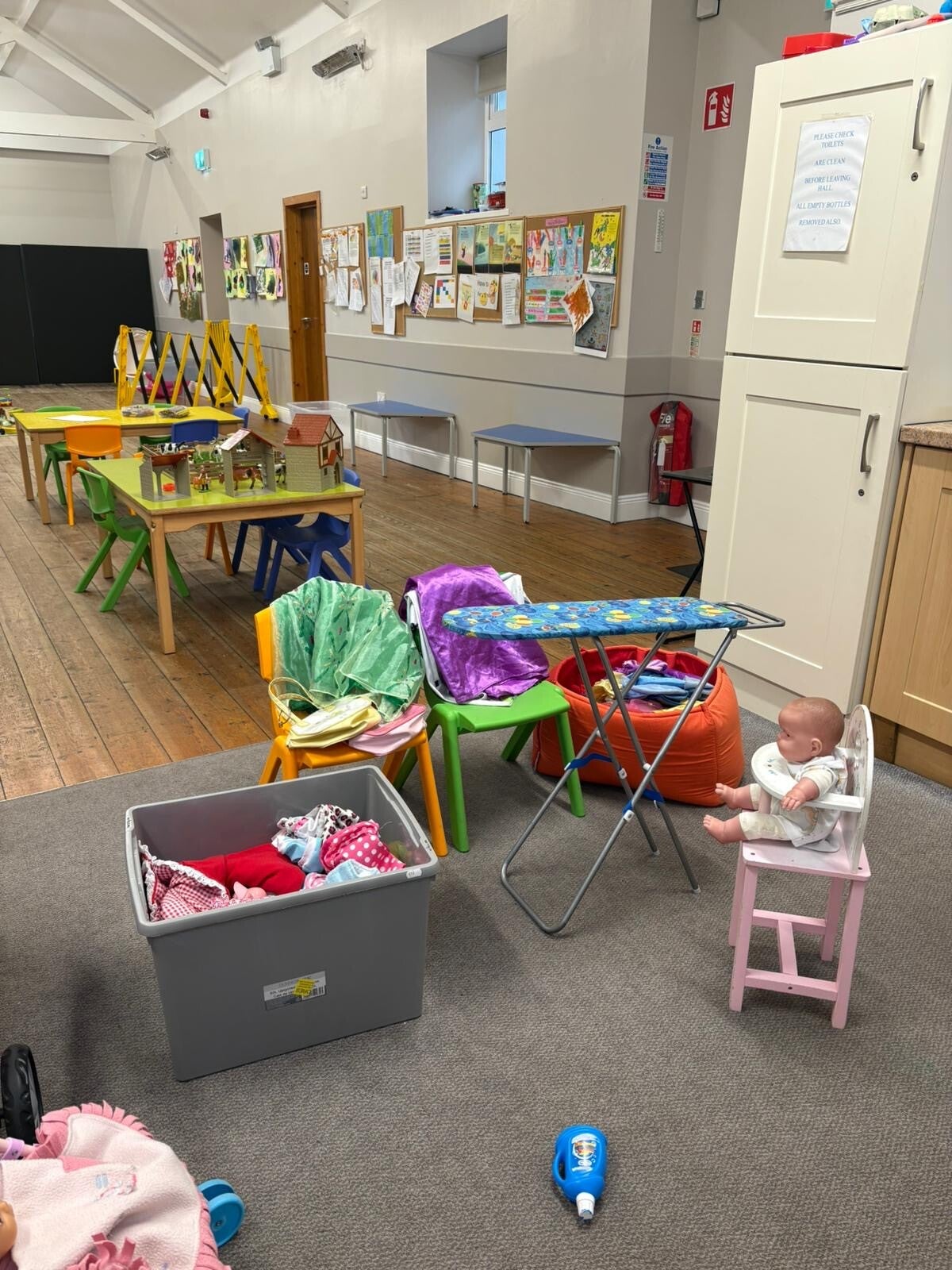A view of Room no.2 where there are tables laid out  with chairs and  an area with a toy ironing board and a basket of washing and a baby in a highchair