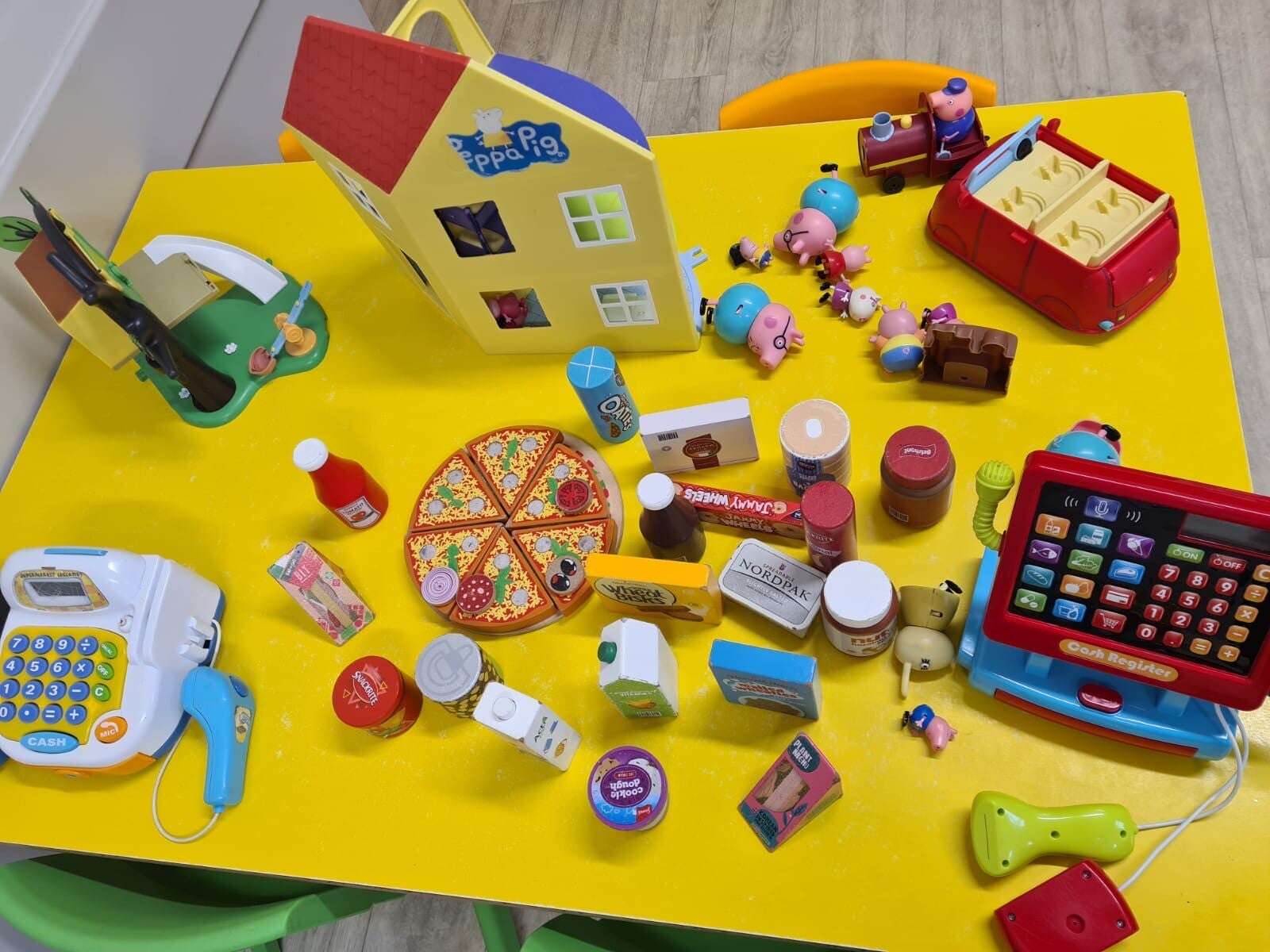 A yellow table displaying Peppa Pig toys, car and house along with a toy cash register and toy food and grocery items.