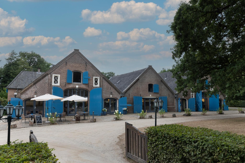 Een sfeervolle boerderij met blauwe luiken, een terras met parasols en een groene omgeving, geschikt voor evenementen en samenzijn.