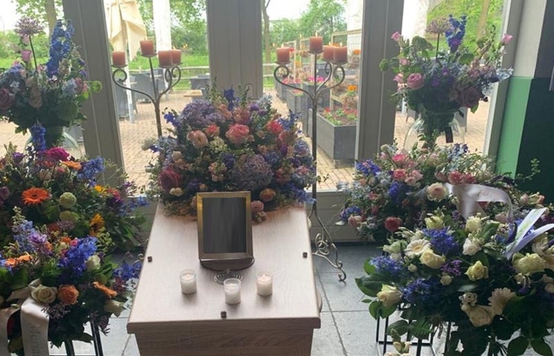 Uitvaartkist omringd door kaarsen standaards en vele boeketten van bloemen met heel veel kleur voor ramen.