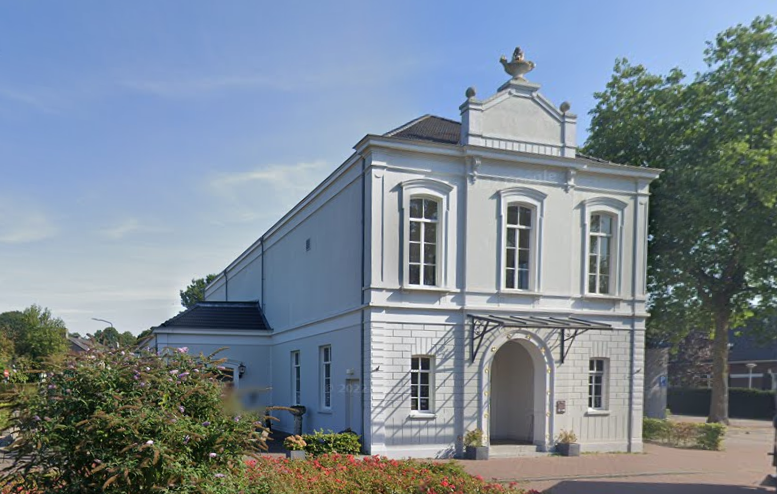 Klassiek wit gebouw met hoge ramen en sierlijke details, omgeven door groen en badend in warm zonlicht