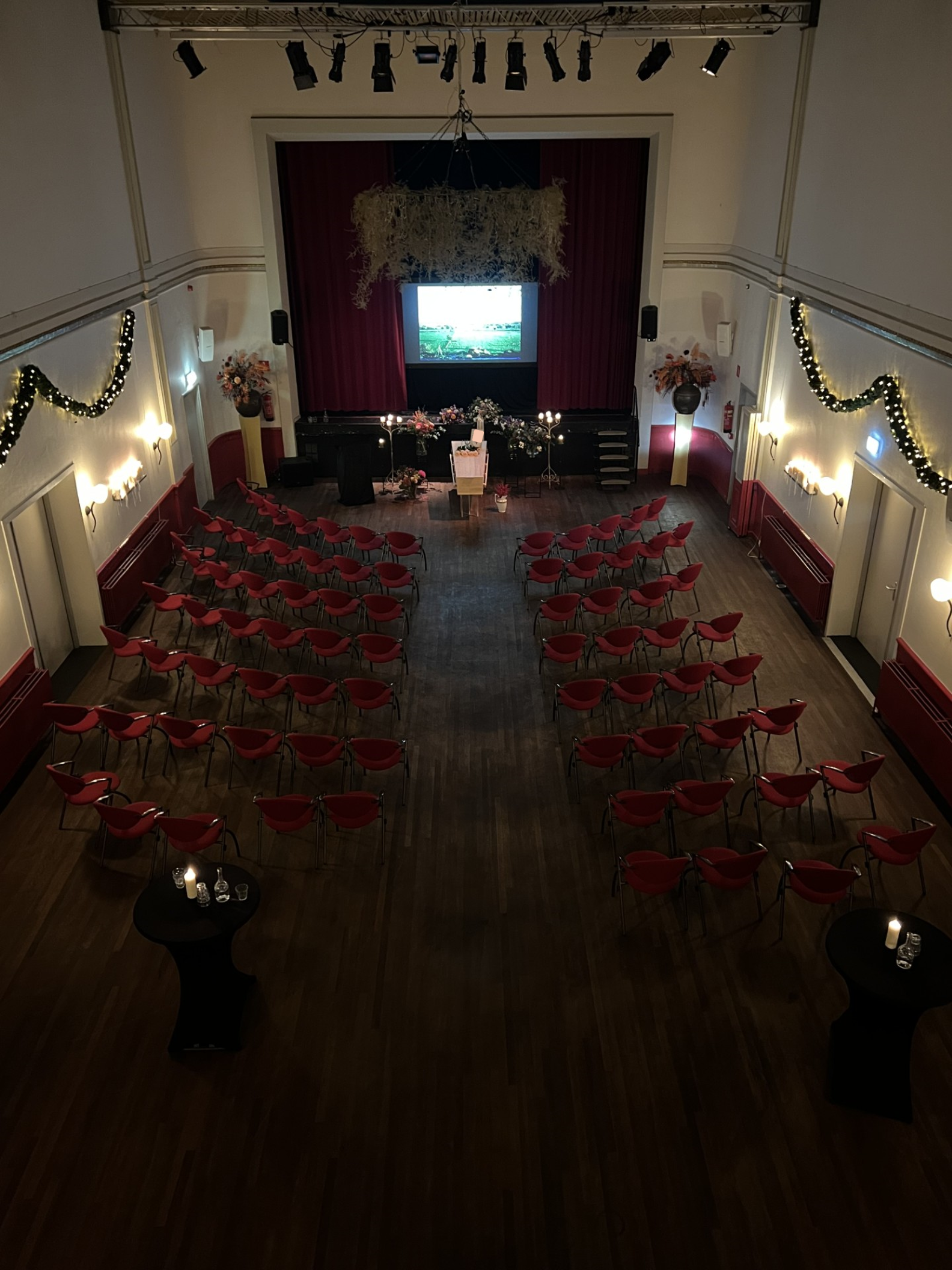 Intieme uitvaartopstelling in de Concertzaal Oosterbeek met roodgordijnen, kaarslicht, bloemen en projectiescherm