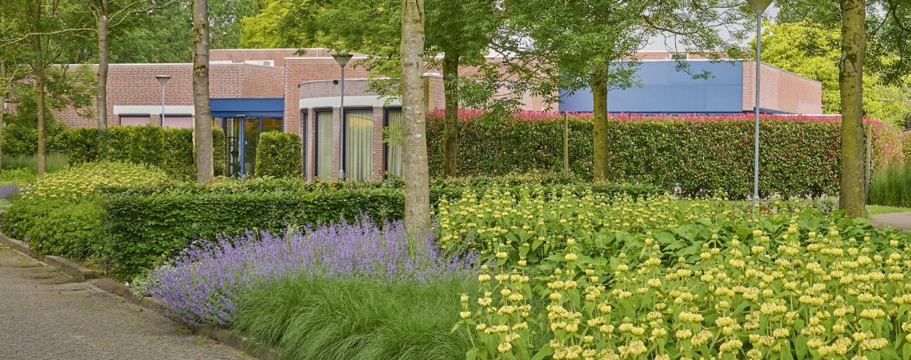 Een rustig uitvaartcentrum met een groene tuin, bloemen en bomen rondom het gebouw, wat bijdraagt aan een serene en rustige sfeer.