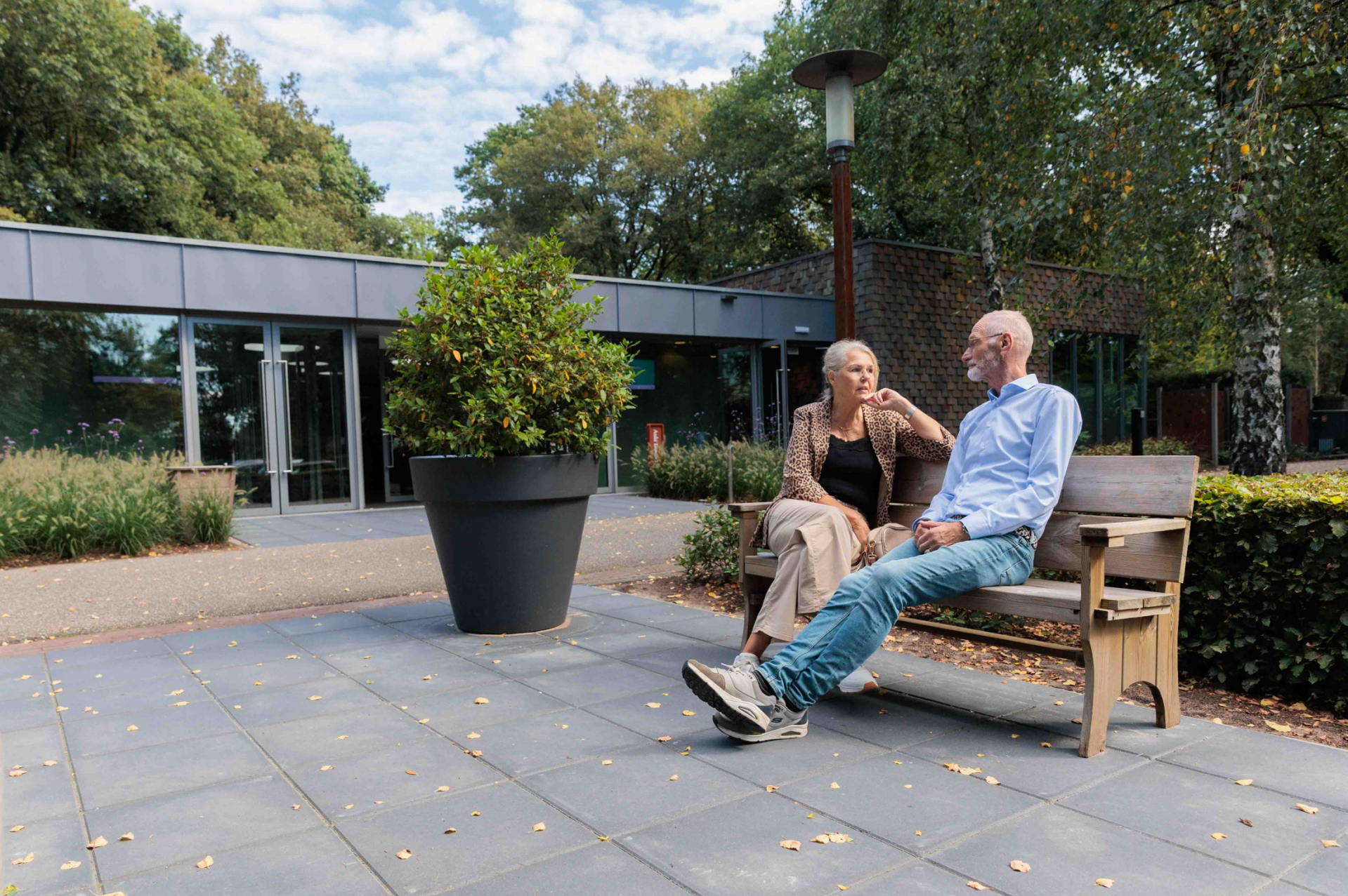 Man en vrouw zitten op een bankje voor een crematorium naast een plant in een bloempot.